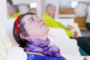 Eine Frau mit geistiger Behinderung schaut an die Decke, ausgestreckt auf einem Schaukelstuhl. Im Hintergrund sehen wir eine weitere Frau auf einem Sofa sitzen