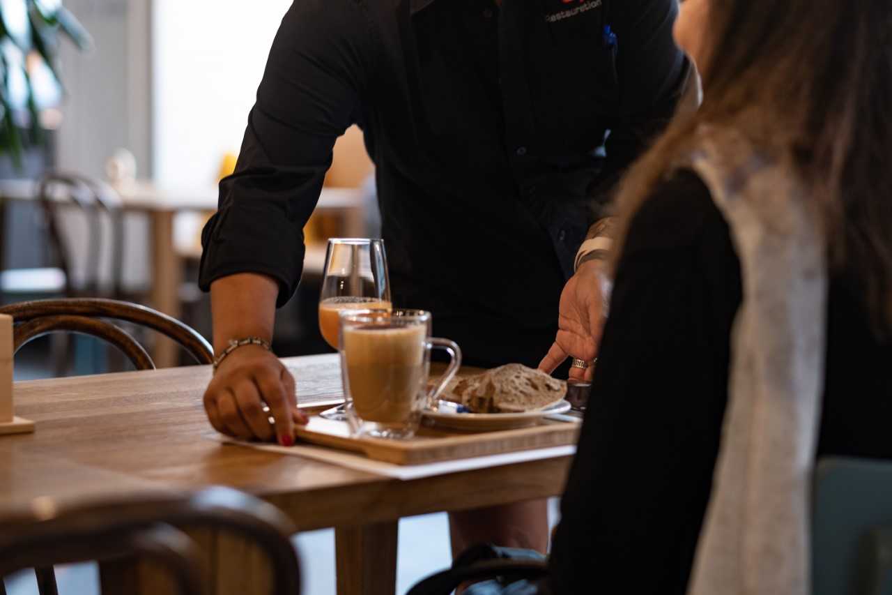 Eine Person serviert einer am Tisch sitzenden Frau einen Kaffee.