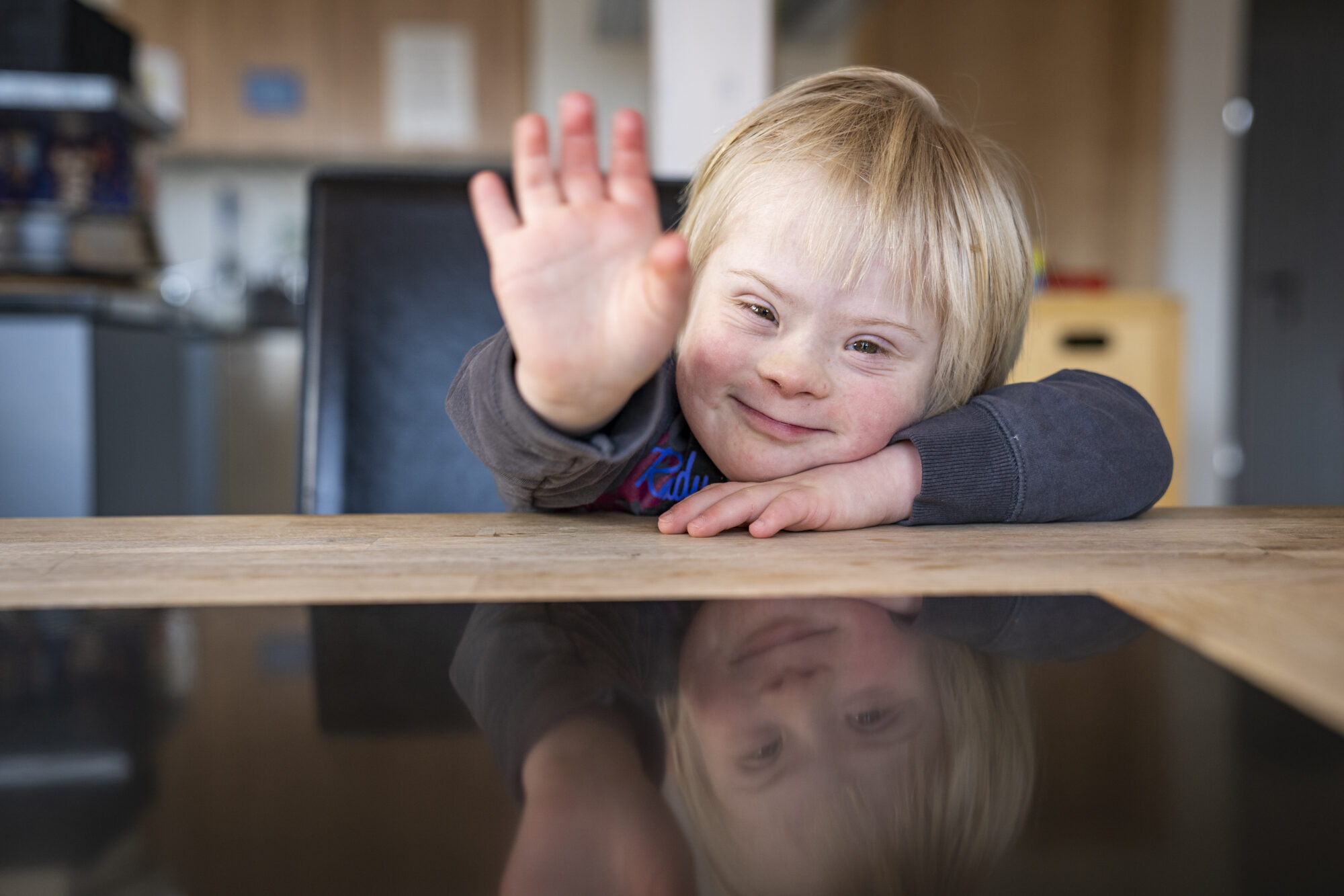Ein Kind mit Trisomie21 sitzt am Esstisch und winkt in die Kamera.