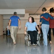 Zwei Frauen gehen, indem sie sich an den Schultern festhalten, während eine Frau im Rollstuhl voranschreitet.