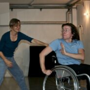 Eine Frau im Rollstuhl macht eine Handbewegung, während eine andere den Rollstuhl zieht.