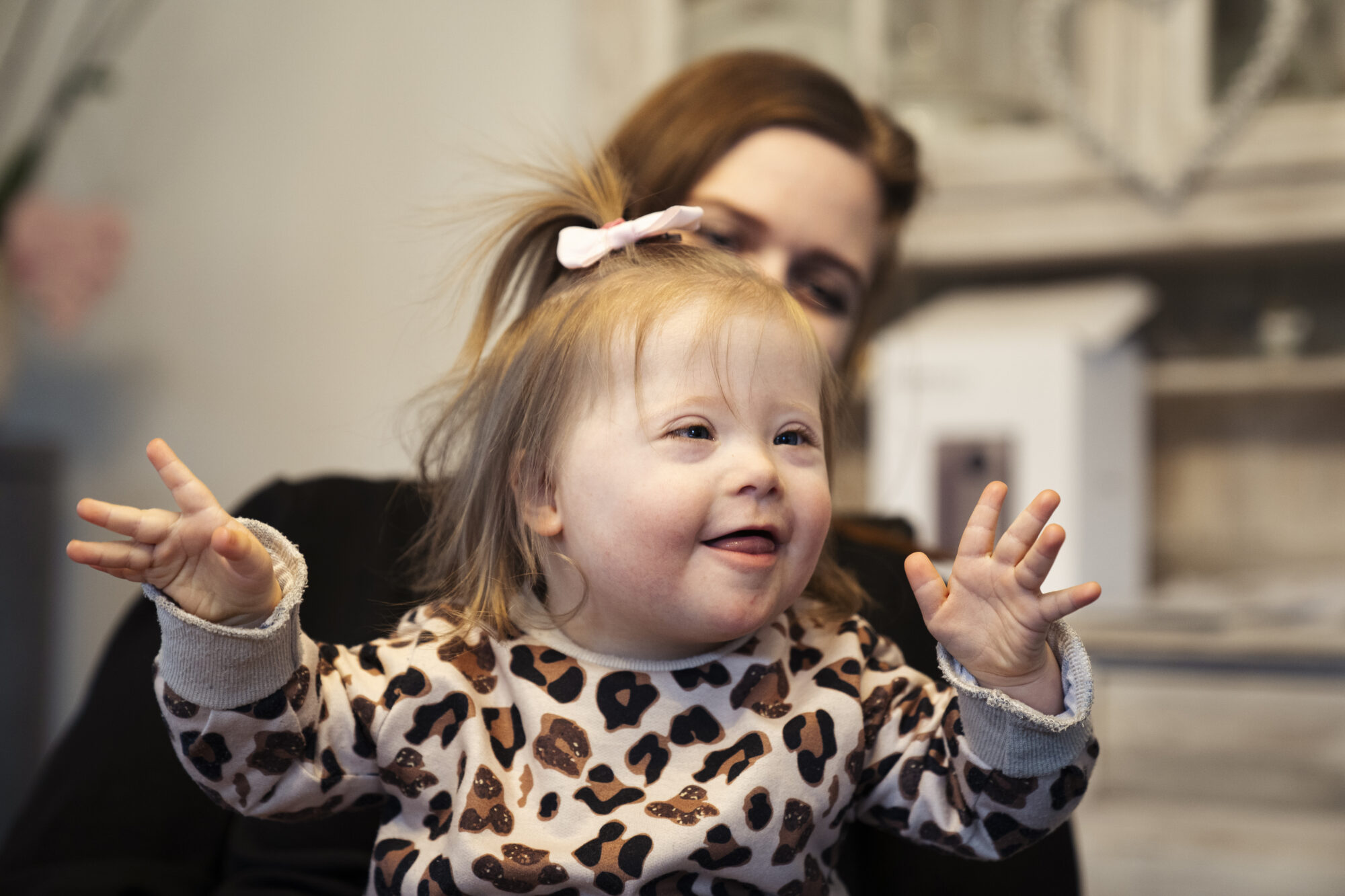 Ein kleines Mädchen mit Trisomie 21 hat die Hände erhoben und lächelt.