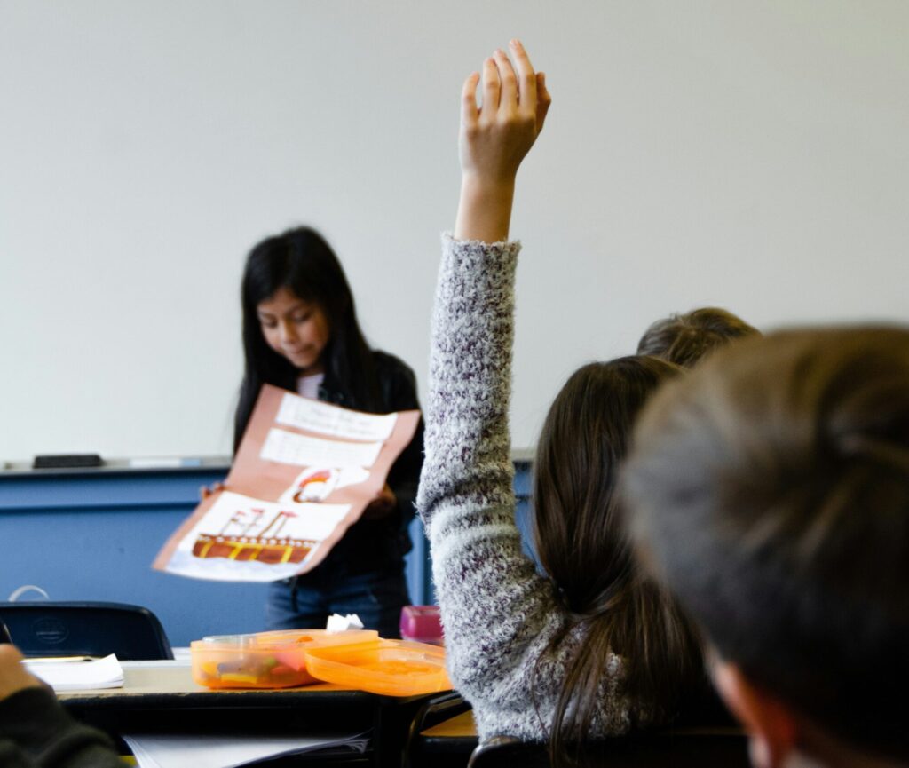In einem Klassenzimmer hebt ein Kind von hinten die Hand.